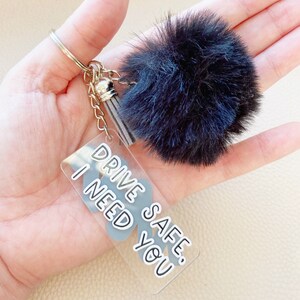 May include: A black and white keychain with a fluffy black pom-pom charm and a clear acrylic charm with the text "Drive Safe. I Need You".