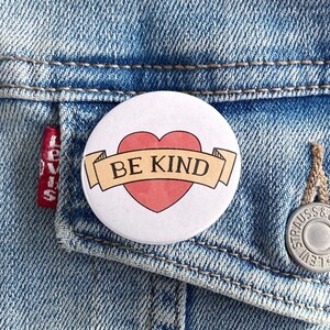 May include: A white button with a red heart and a yellow banner that says "BE KIND".