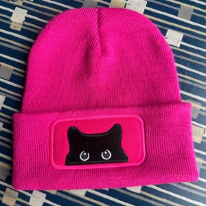 May include: A vibrant pink beanie hat with a black cat face patch. The patch shows a black cat silhouette with white eye details. The hat is made of a knitted material and has a folded brim.