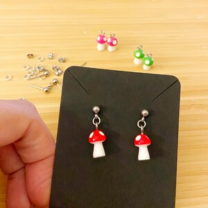 May include: A pair of silver stud earrings with red and white mushroom charms. The earrings are displayed on a black card.