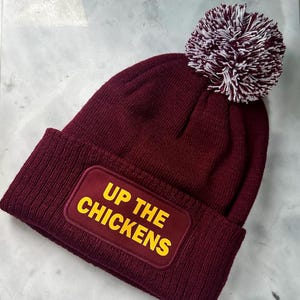 May include: A maroon knitted beanie hat with a white and maroon pom-pom. The hat has a maroon patch with the words "UP THE CHICKENS" in yellow. The beanie is made of a soft, knitted material.