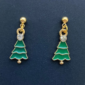 May include: A pair of Christmas tree earrings. Each earring has a green enamel tree with gold-coloured trim and a clear crystal accent. The earrings have a gold-coloured ball stud and a small loop connecting the tree charm.