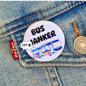 May include: A white button with a blue and pink bus illustration and the text "BUS WANKER" in black. The button has a white speech bubble with the text "!?!?*" in black.