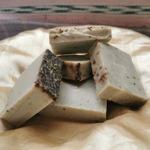 May include: A close-up of five bars of handmade soap. The soap is a light green color with brown flecks throughout. The soap is arranged in a pile on a gold fabric surface.