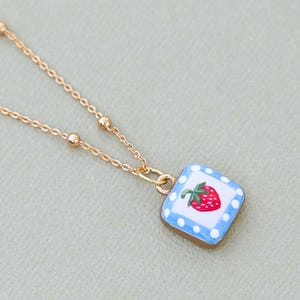 May include: A gold-colored necklace with a square pendant featuring a red strawberry illustration. The pendant has a light blue border with white polka dots. The necklace chain has small, round beads.