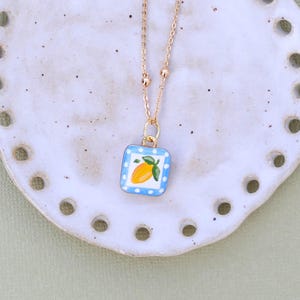 May include: A gold-colored necklace with a square pendant featuring a lemon design. The pendant has a blue and white checkered border. The necklace is displayed on a white, round plate with evenly spaced holes around the edge.