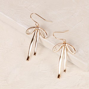 May include: A pair of gold-toned dangle earrings with a bow design. The earrings are on a white surface.