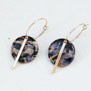 May include: A pair of hoop earrings. Each earring features a circular, marbled pendant in shades of black, brown, and purple, accented with a gold-colored bar. The earrings have gold-colored hoops.