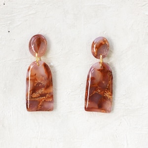 May include: A pair of dangle earrings with a marbled, reddish-brown and gold pattern. The earrings have a rounded top and a rectangular bottom with a curved arch. The earrings are connected with gold-colored hardware.