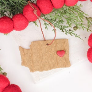 May include: A tan-colored ornament shaped like the state of Washington, with a red heart and a cable knit design. The ornament hangs from a brown string and is surrounded by red felt ball garland and evergreen branches.
