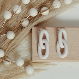 May include: A pair of white dangle earrings featuring a double-link chain design. Each earring consists of two interlocking oval-shaped links with a subtle speckled texture throughout the matte white surface, attached to a simple metal post backing.
