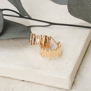 May include: Gold hoop earrings with a geometric design. The earrings are made of a series of vertical bars that are connected together.
