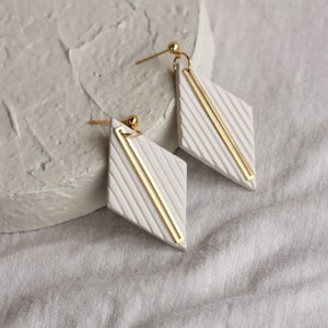 May include: A pair of white diamond-shaped earrings with gold accents. The earrings have a textured surface and are accented with two gold bars.