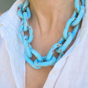 May include: A chunky, light blue chain necklace with distressed brown accents. The necklace is worn over a white collared shirt. The chain links are large and oval-shaped, creating a bold statement piece.