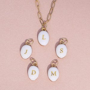 May include: A gold-colored chain necklace with a white oval pendant featuring the letter "L". Four additional white oval pendants with the letters "J", "S", "D", and "M" are also visible. The pendants have gold-colored accents and are set against a pink background.