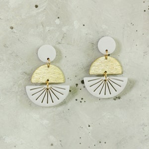 May include: A pair of white and gold geometric earrings. The earrings are made of polymer clay and feature a sunburst design.