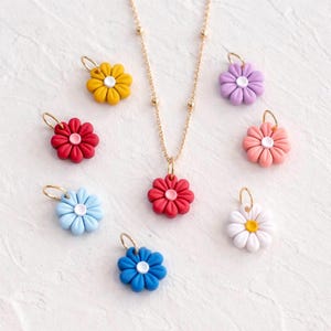 May include: A gold-colored necklace with a red flower pendant, surrounded by eight colorful flower charms. The charms are in various colors, including yellow, red, blue, purple, pink, and white. Each flower has a gold-colored loop.