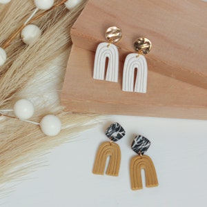 May include: Two pairs of handmade polymer clay earrings. One pair features white rainbow arches with gold textured tops. The other pair features mustard yellow rainbow arches with black and white marbled tops.