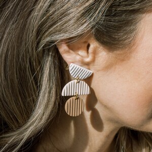 May include: A pair of dangle earrings with three geometric shapes. The top shape is a white half-circle with a textured pattern. The middle shape is a white half-circle with a textured pattern. The bottom shape is a brown circle with a textured pattern.
