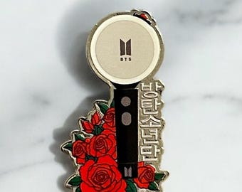May include: A silver metal pin featuring a stylized black and white image of a BTS light stick with red roses surrounding the base. The light stick has the BTS logo and the text "BTS" on the top. The text "하이터스턴트" is written on the side of the light stick.