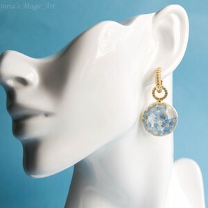 Puede incluir: Aretes de aro dorados con un dije redondo de resina transparente lleno de flores azules de nomeolvides.