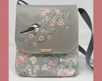 Puede incluir: Un bolso bandolera gris con estampado floral y un pájaro bordado en una rama. El bolso tiene cierre de solapa y una correa negra. El nombre de la marca "THREADGEAR" está impreso en una etiqueta metálica.