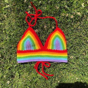 May include: A handmade crochet halter top featuring a vibrant rainbow pattern with horizontal stripes of red, orange, yellow, green, blue, and purple. The garment includes thin red yarn ties at the neck and base for an adjustable fit.