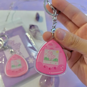 May include: Pink and clear plastic keychain shaped like a Tamagotchi with a cartoon llama on the screen. The keychain has a silver metal ring.