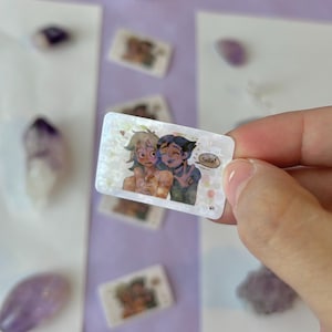 May include: A rectangular sticker with an illustration of two cartoon characters hugging, with one saying "Smile!" in a speech bubble. The sticker has a holographic, iridescent finish and is held by a hand.