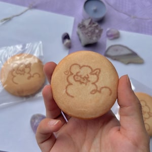 Pode incluir: Dois biscoitos redondos de cor castanho claro com um contorno castanho de uma ovelha de desenho animado em cada um. A ovelha tem um coração acima da cabeça e está rodeada por uma nuvem.