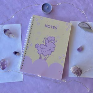 Può includere: Un quaderno a spirale viola e giallo con un lama di cartone animato sulla copertina. Il quaderno dice "NOTES" sulla parte anteriore. Il quaderno è circondato da cristalli viola e una ghirlanda di luci.
