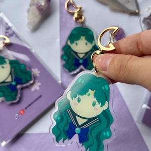 Può includere: Portachiavi in acrilico trasparente con un personaggio dei cartoni animati con capelli verde acqua, un fiocco blu e un'uniforme da marinaio. Il portachiavi ha una chiusura color oro. Lo sfondo include altri portachiavi e confezioni viola.