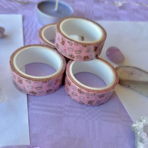 以下が含まれることがあります： 茶色のクマとピンクのハートのパターンが描かれたピンクの和紙テープが4つ巻かれています。テープは紫色の表面に積み重ねられています。
