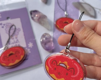 May include: Clear acrylic charm featuring a red donut design with a cute face and glitter accents. The charm has a black lanyard attached. The background includes crystals and a purple package.