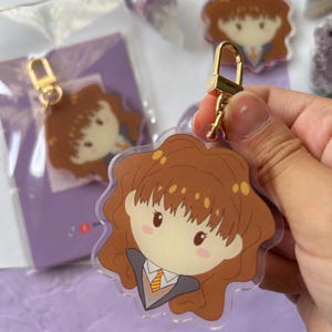 Può includere: Un portachiavi in acrilico trasparente con un'illustrazione a cartoni animati di un personaggio con i capelli castani, che indossa un blazer grigio e una cravatta a righe. Il portachiavi ha una chiusura color oro. Altri portachiavi sono visibili sullo sfondo.