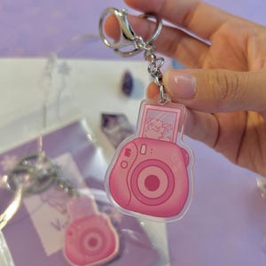 May include: Pink and white camera keychain with a small image of a cartoon bear inside the viewfinder. The keychain is made of clear acrylic and has a silver metal key ring.