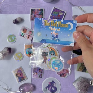 Puede incluir: Una bolsa de plástico transparente que contiene un juego de pegatinas con el texto "The Owl House" y "Sticker set x 14 pcs". Las pegatinas son de varias formas y tamaños, con ilustraciones coloridas. El fondo es una superficie de color púrpura claro con otros elementos decorativos.