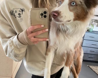 May include: A woman wearing a beige sweatshirt with a black and white dog illustration and the word "JIM" printed on it. She is holding a brown and white dog with blue eyes.