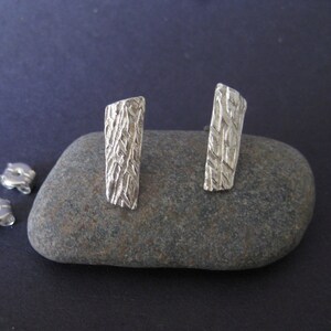 Puede incluir: Un par de pendientes de plata con un diseño rectangular texturizado. Los pendientes se muestran sobre una roca gris.