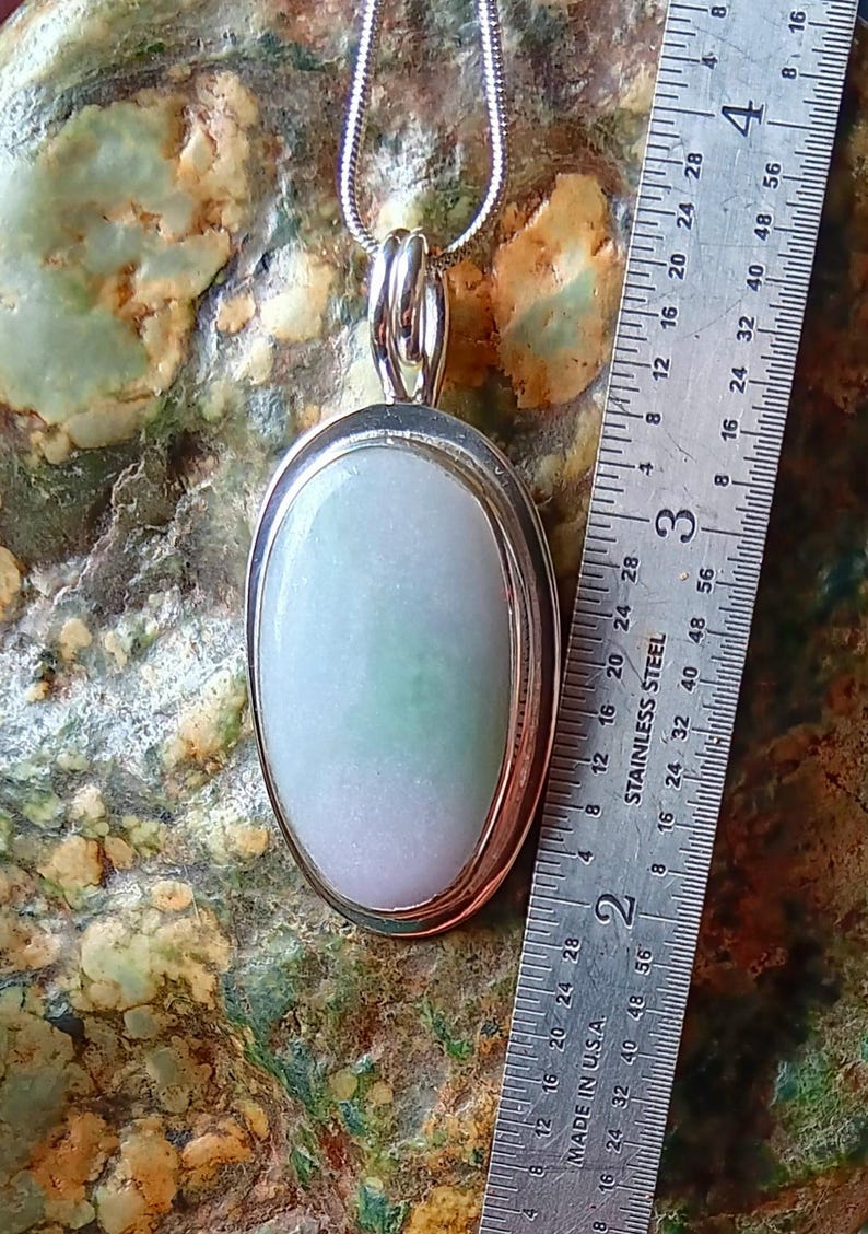 Op de afbeelding: Ovale hanger met een lichtgroene en lavendelkleurige steen gezet in een zilverkleurig metalen frame. De hanger hangt aan een zilveren ketting. Een liniaal toont dat de hanger ongeveer 3,8 cm lang is.