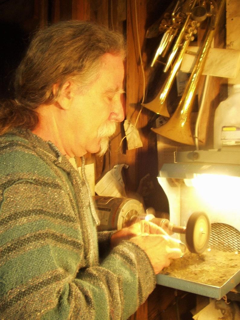May include: A person wearing a teal and grey striped sweater using a motorized buffing machine to polish a small metal object. The workspace features a bright lamp and brass musical instruments hanging on the wooden wall in the background.