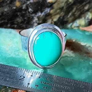 May include: A silver ring with a large, oval, bright turquoise stone. The ring is set in a bezel setting. A stainless steel ruler is in the foreground, showing measurements in inches.