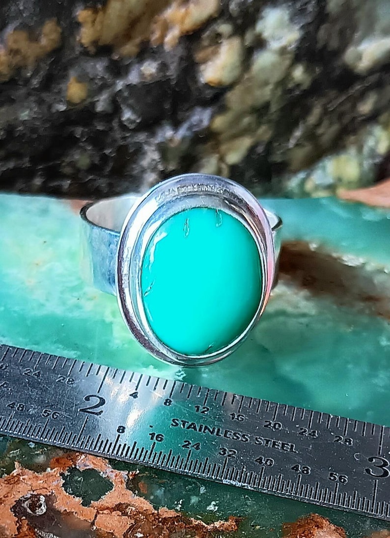 May include: A silver ring with a large, oval, bright turquoise stone. The ring is set in a bezel setting. A stainless steel ruler is in the foreground, showing measurements in inches.