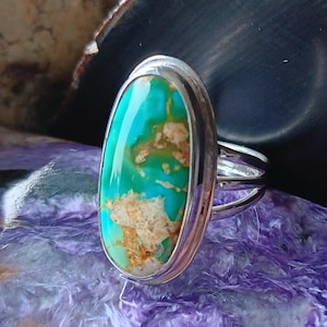 May include: A silver ring featuring a large, oval-shaped turquoise stone with vibrant teal and green hues and prominent tan matrix inclusions. The stone is set in a smooth, polished silver bezel with a simple, split-shank silver band design.