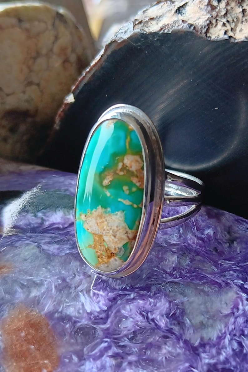 May include: A silver ring featuring a large, oval-shaped turquoise stone with vibrant teal and green hues and prominent tan matrix inclusions. The stone is set in a smooth, polished silver bezel with a simple, split-shank silver band design.