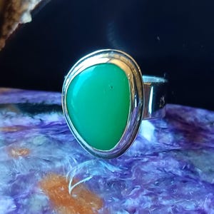 May include: A silver ring featuring a smooth, asymmetrical, vibrant green cabochon stone set in a polished metal bezel. The ring band is made of a plain, reflective silver-toned metal that curves around the finger, showcasing a clean design.