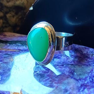May include: A silver ring featuring a smooth, teardrop-shaped green gemstone set in a polished metal bezel. The band is wide and reflective, showcasing a metallic sheen that complements the vibrant, solid green color of the central stone.