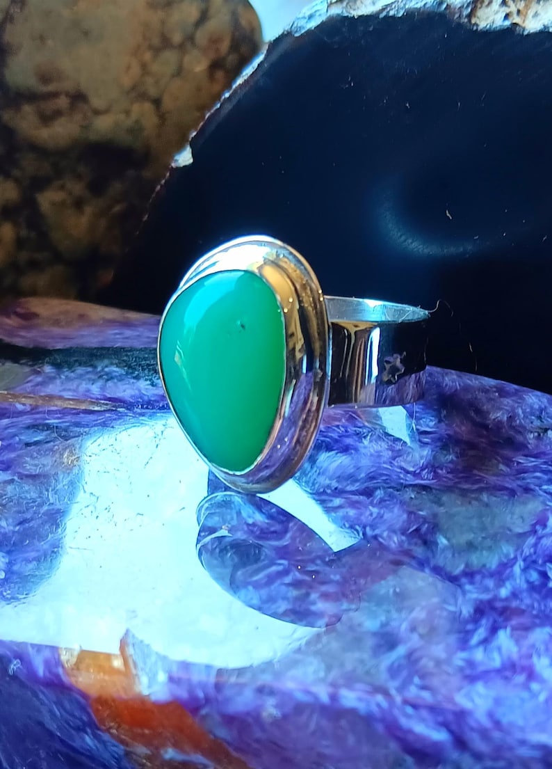 May include: A silver ring featuring a smooth, teardrop-shaped green gemstone set in a polished metal bezel. The band is wide and reflective, showcasing a metallic sheen that complements the vibrant, solid green color of the central stone.