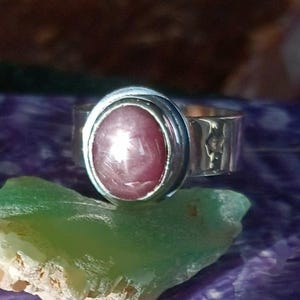 May include: A silver ring featuring an oval, pink gemstone. The ring's band has a textured design. The gemstone has a star-like effect. The ring is displayed on a green and purple surface.