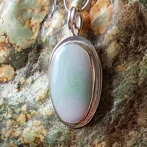 Op de afbeelding: Ovale jade hanger aan een zilveren ketting. De jade steen heeft een lichtgroene en witte kleur. De hanger is gezet in een zilveren bezel. De ketting is een dunne, zilveren slangenketting. De achtergrond is een getextureerde, groen-bruine steen.